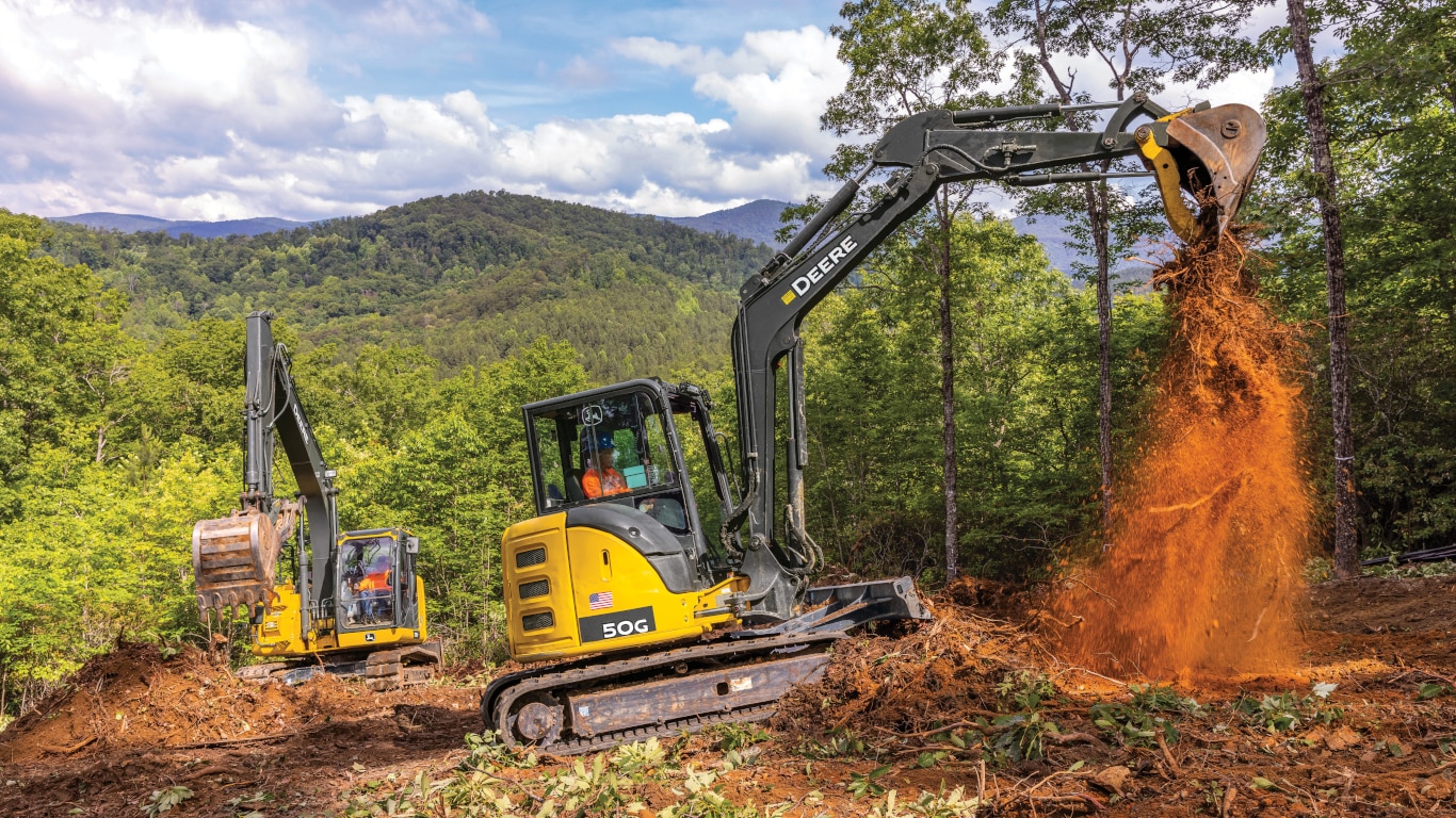 Two John Deere 50G Excavators scooping dirt in a forest with mountains in the background.